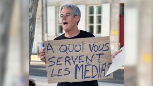 Un quinquagénaire aux cheveux courts argentés se tient debout dans une rue. Il tient devant sa poitrine une pancarte en carton, sur laquelle il est écrit en grosses lettres "A quoi vous servent les médias". Il a la tête légèrement tournée vers sa droite, la bouche ouverte. Il semble alpaguer quelqu'un à distance.