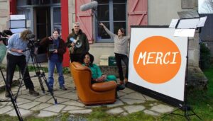 Cinq personnes se tiennent devant une maison aux volets rouges. Une sixième les filme avec une caméra posée sur un trépied. Parmi les 5 personnes, l'une d'elles est penchée sur sa propre caméra. eux autres tiennent avec fierté un clap de cinéma et un mégaphone. Une autre brandit au-dessus de leurs têtes une perche de micro. La dernière est installée dans un fauteuil en cuir, posée à même le pavement de la devanture de la maison. Elle nous adresse un grand sourire. Face au canapé, au écran de home-cinéma est déployé, sur lequel on peut lire en grand "merci".