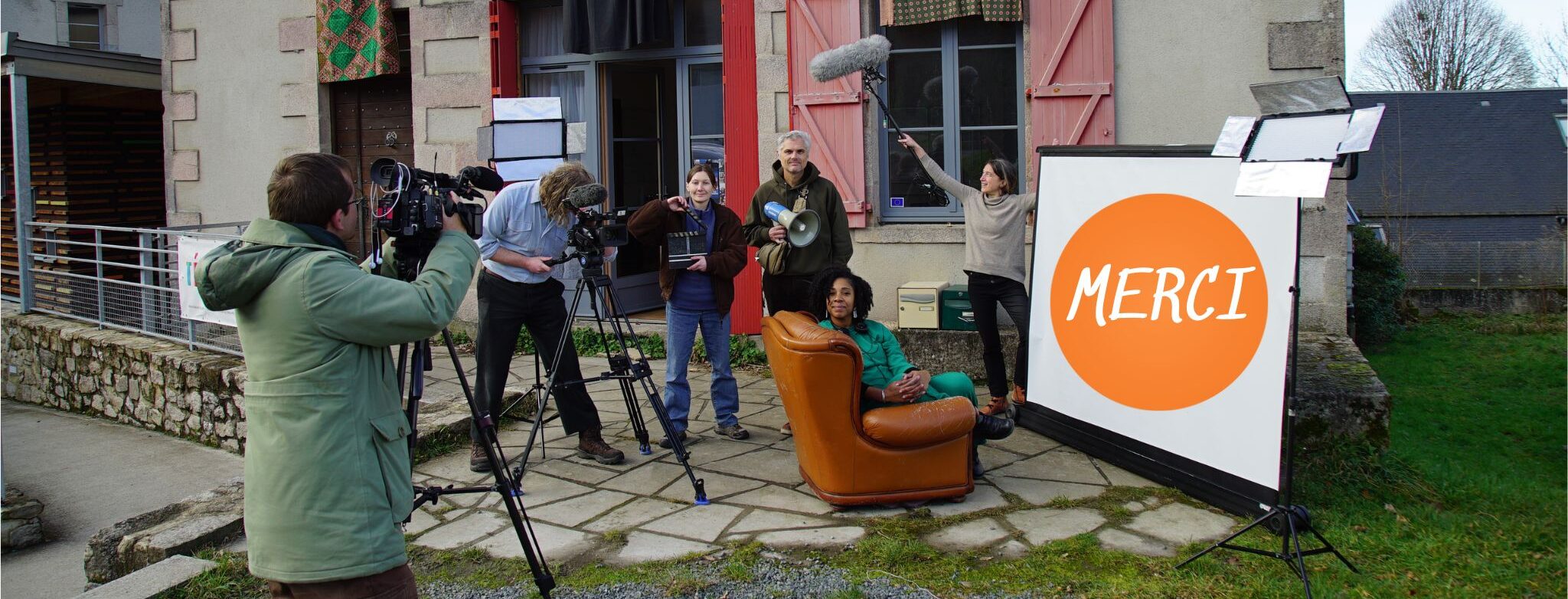 Cinq personnes se tiennent devant une maison aux volets rouges. Une sixième les filme avec une caméra posée sur un trépied.
Parmi les 5 personnes, l'une d'elles est penchée sur sa propre caméra. eux autres tiennent avec fierté un clap de cinéma et un mégaphone. Une autre brandit au-dessus de leurs têtes une perche de micro. La dernière est installée dans un fauteuil en cuir, posée à même le pavement de la devanture de la maison. Elle nous adresse un grand sourire. Face au canapé, au écran de home-cinéma est déployé, sur lequel on peut lire en grand "merci".