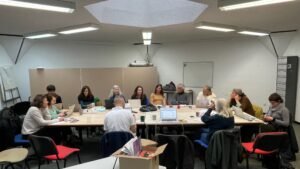 13 personnes sont réunies autour d'une grande table rectangulaire, dans une salle de réunion. La plupart ont un ordinateur portable devant eux. Ils s'écoutent, certains consultent leurs notes. Posé devant eux sur une table à part, un carton ouvert. Il en dépasse des brochures d'informations sérigraphiées.