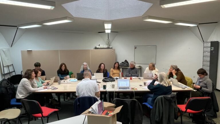 13 personnes sont réunies autour d'une grande table rectangulaire, dans une salle de réunion. La plupart ont un ordinateur portable devant eux. Ils s'écoutent, certains consultent leurs notes. Posé devant eux sur une table à part, un carton ouvert. Il en dépasse des brochures d'informations sérigraphiées.