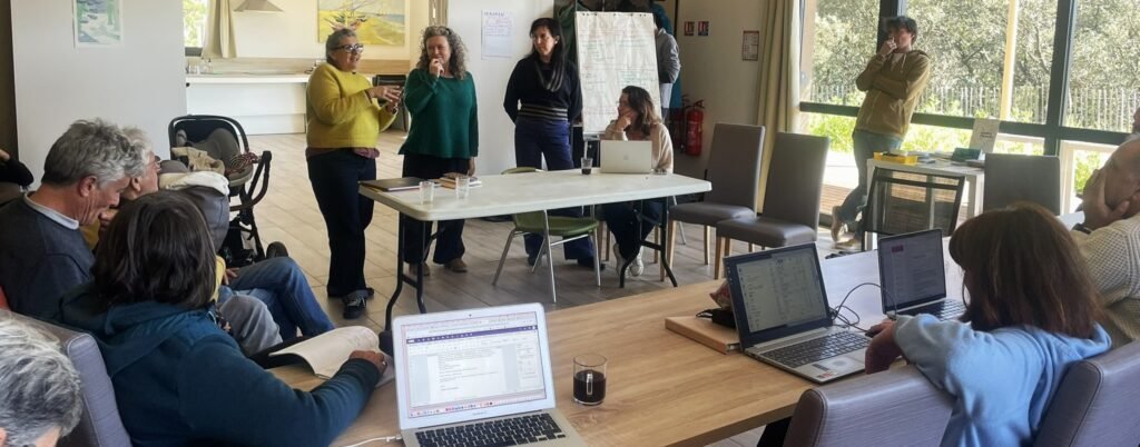 Dans une salle de réunion donnant sur une baie vitrée, les contributrices et contributeurs de la Fédération de l'audiovisuel participatif sont répartis autour d'un rectangle de tables. 2 femmes debouts s'adressent au reste du groupe.